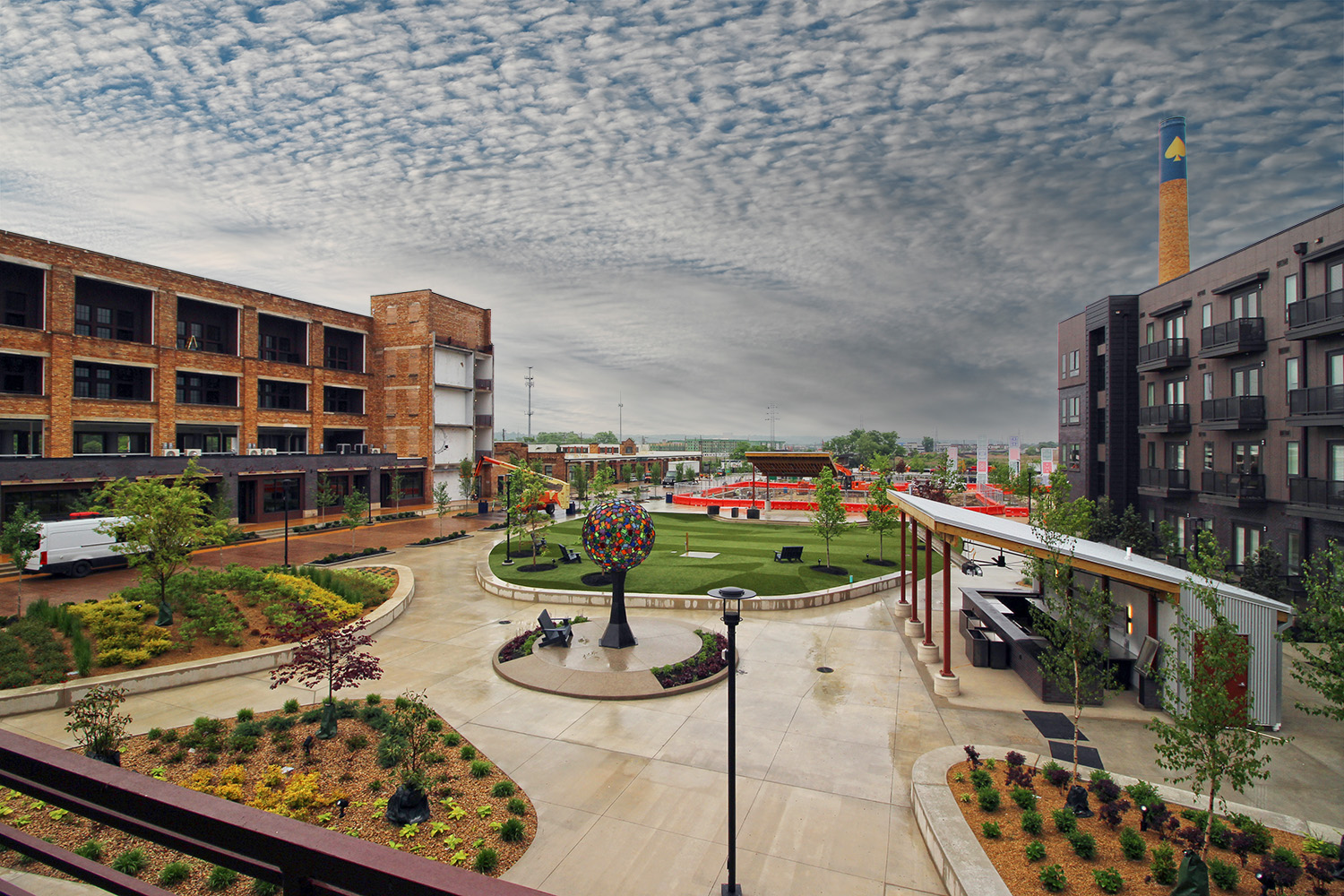Image of a public square with a sculpture in the middle surrounded by brick buildings, landscaped planter beds, and a smokestack in the background.